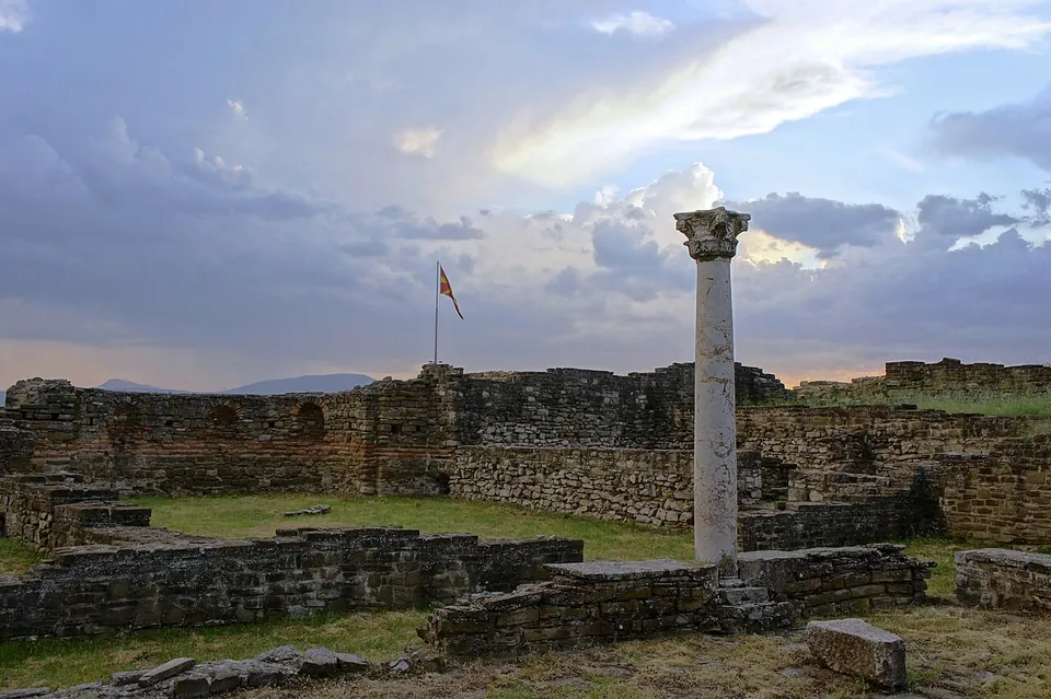 A single marble column with a carved capital standing among stone walls under an evening sky, a flag flying behind.