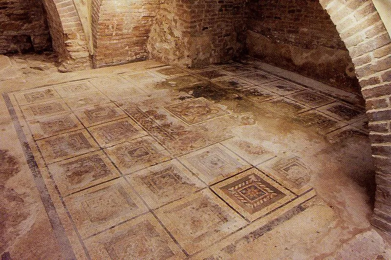 A floor mosaic laid out as a grid of square panels in red, ochre and dark tones framed by bands, in a low room with brick walls and a brick vault.