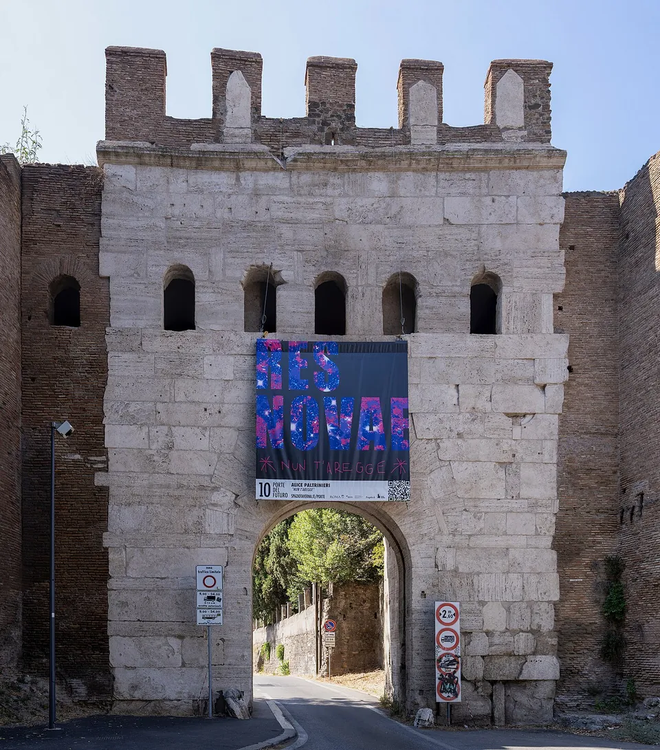 A gate of large pale stone blocks with five small arched windows above a single arched passage and crenellations on top, a banner hanging over the arch and traffic signs at its foot, brick wall on either side.