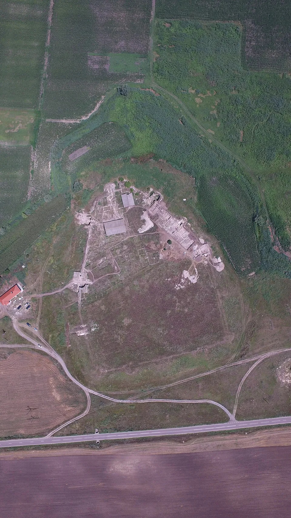 A vertical aerial view of a low rise between green fields and marsh, with the excavated walls and roofed shelters of a fortress on its upper part and a road and tracks curving below.