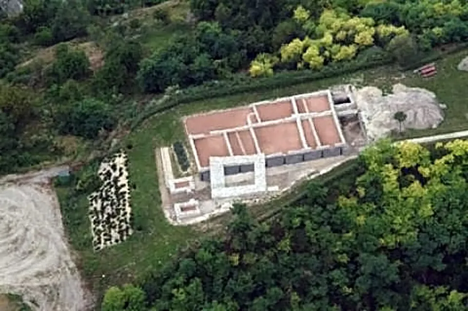 An aerial view of a rectangular building outline of conserved walls divided into rooms floored with reddish gravel, on a clearing surrounded by woodland.