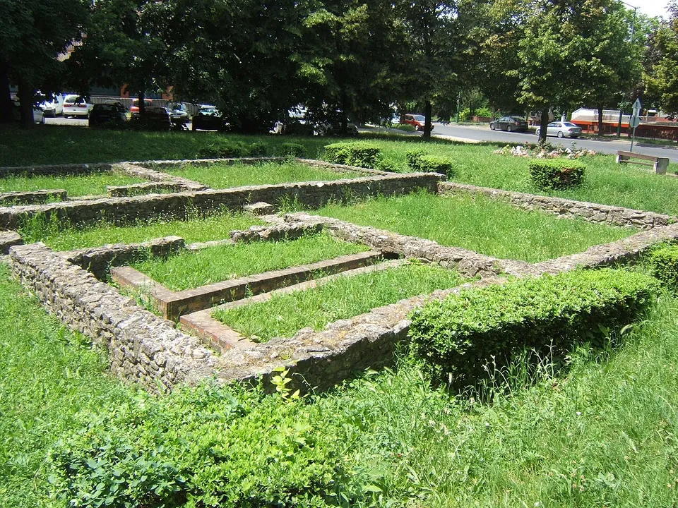 Low stone foundation walls dividing a lawn into rooms, with clipped hedges along one side and a road with parked cars behind.