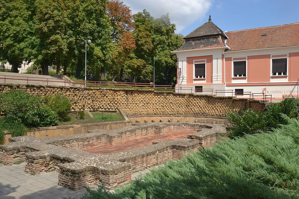 Low foundation walls of brick and stone with curved recesses in a sunken courtyard, below a wall of stone blocks and a pink building with a grey roof.