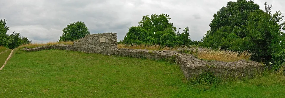 A long, low run of rough stone wall across a grassy slope, rising at one end to a squared block of masonry with a small plaque on it, trees and dry grass behind.