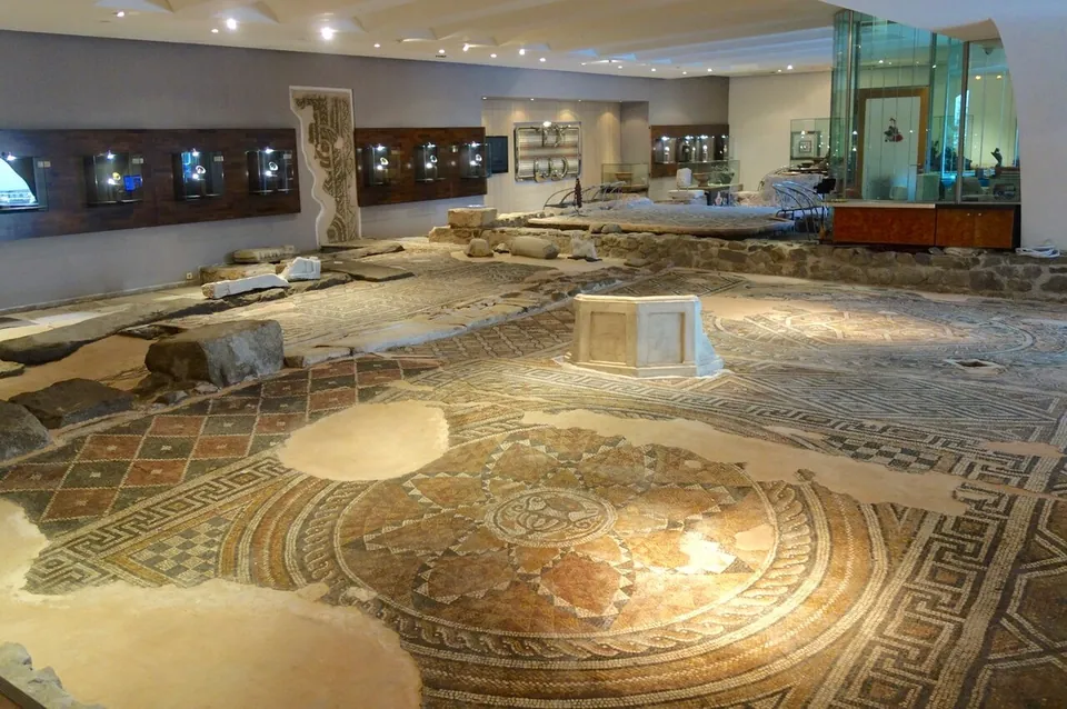 A large expanse of Roman floor mosaic in an indoor hall, with circular and geometric patterns in red, ochre and grey, an octagonal marble basin standing on it, low ruined walls, and display cases along the walls behind.