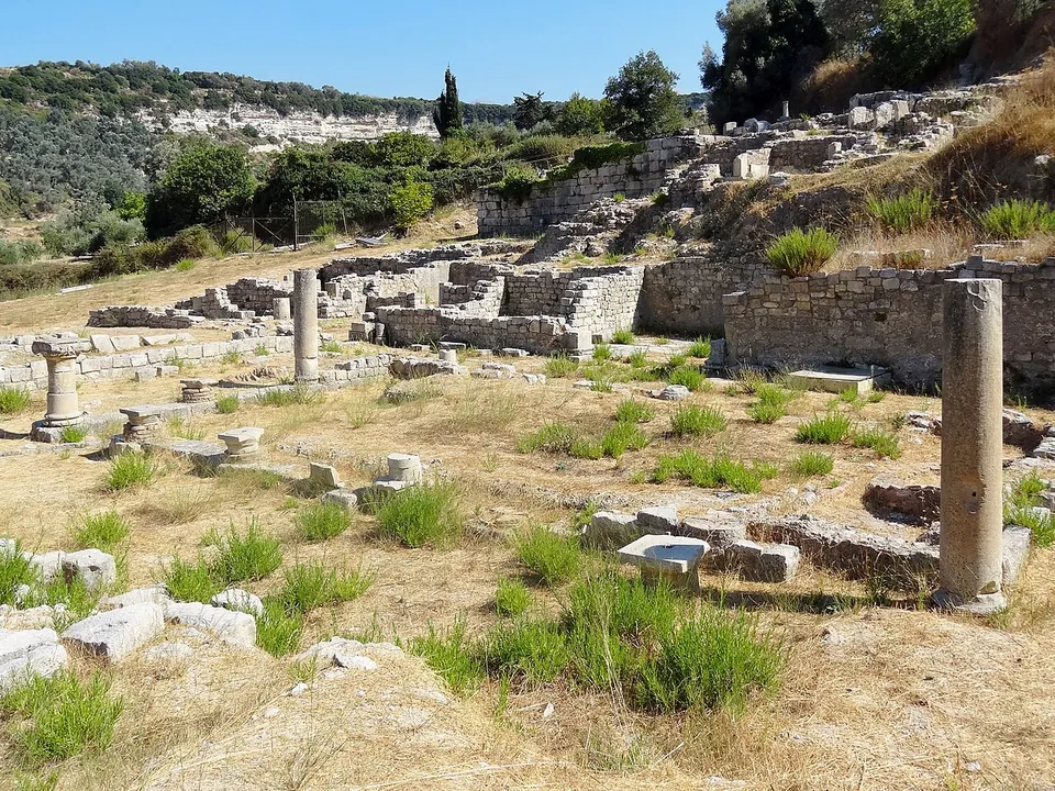 An excavated hillside of low stone walls, column bases and two standing column shafts on dry grass, with terraced walls rising behind and a wooded ridge beyond.
