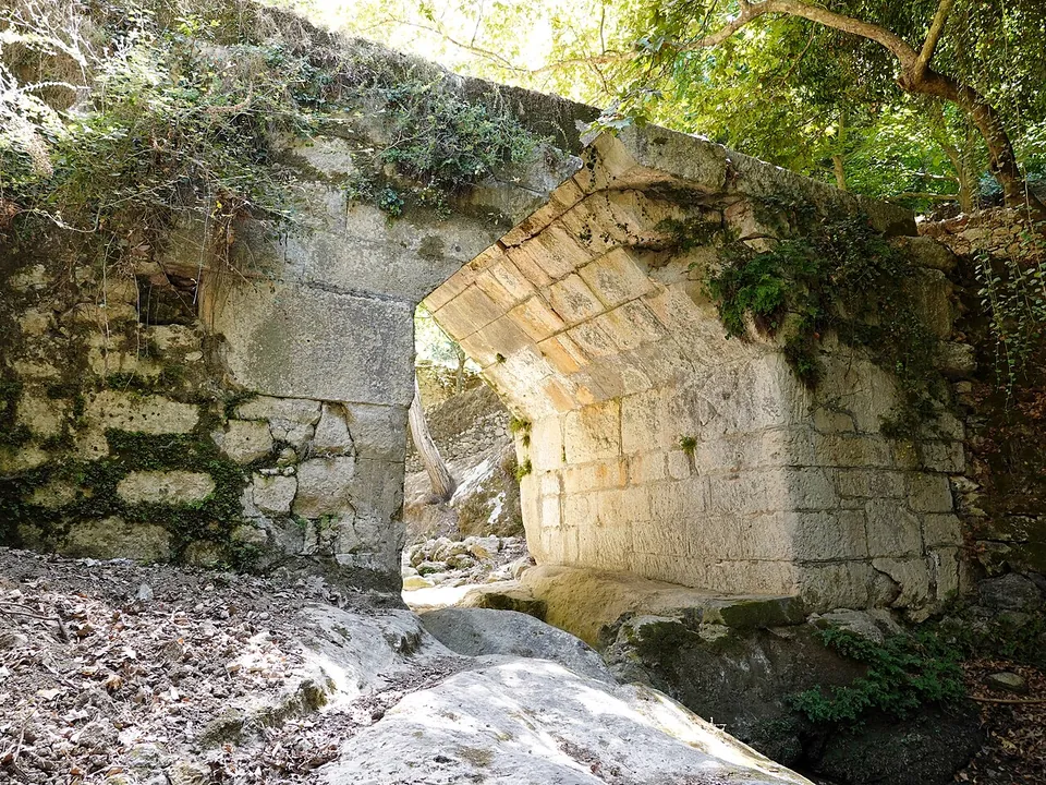 A small bridge of large squared stones over a rocky stream bed, its opening a pointed arch of corbelled blocks, overgrown with ivy and shaded by trees.