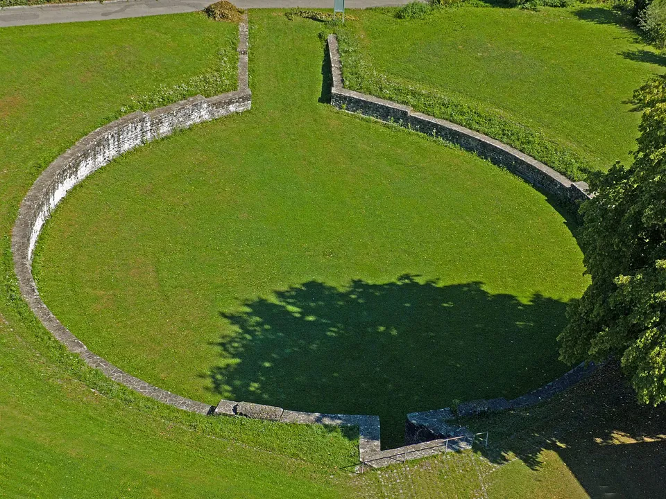 An aerial view of a round grassy arena outlined by a low stone wall, with straight walls marking an entrance at the bottom and the shadow of a tree across it.