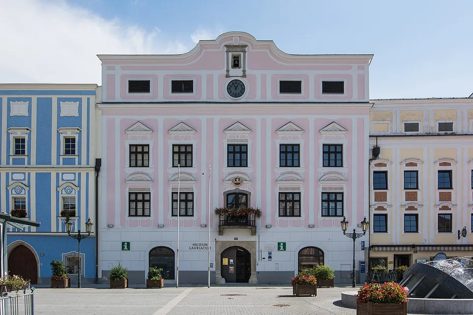 A pink-and-white baroque town house with a clock in its gable and a balcony over the central doorway, on a paved square with flower boxes, between two other painted houses.