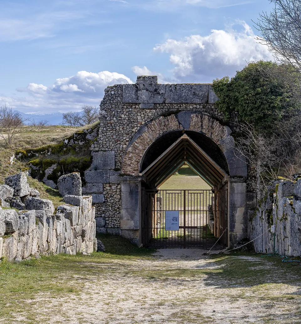 A round arch of brick set in a wall of small rubble stones and large squared blocks, closed by a modern iron gate under a timber shelter, with walls of large irregular stone blocks on either side of a gravel path.