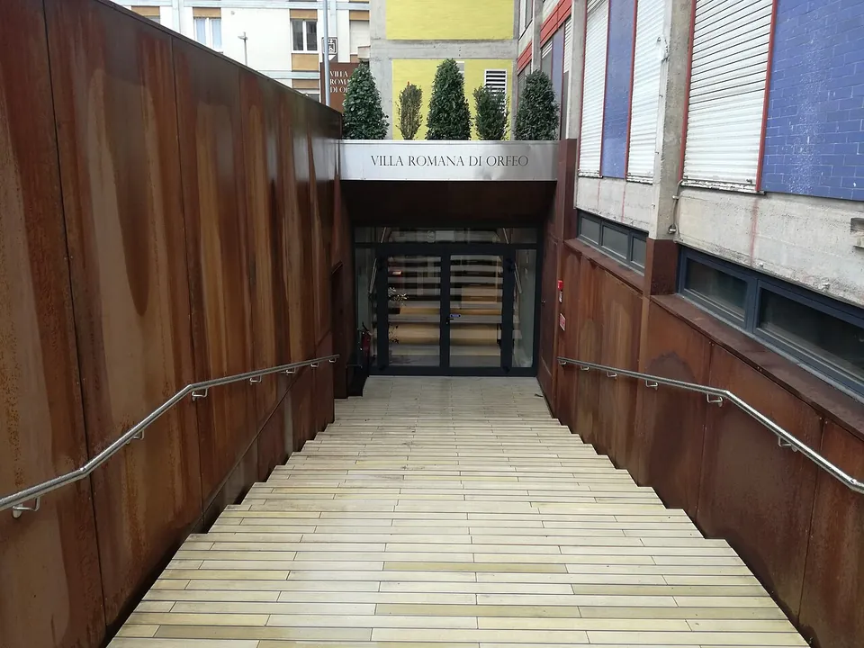 A broad flight of pale stone steps leading down between rust-coloured metal walls to glass doors under a sign reading "Villa Romana di Orfeo".