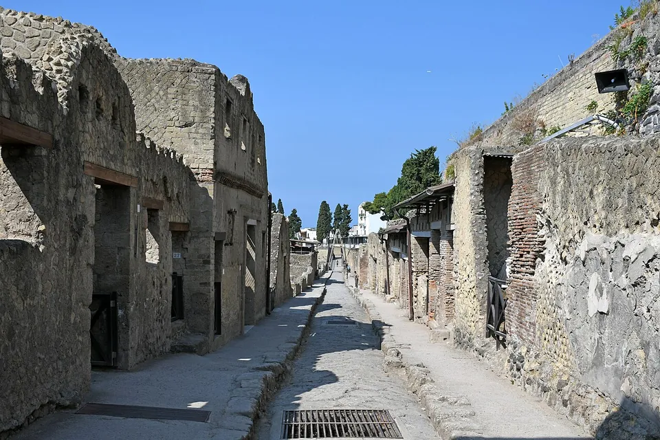 A narrow paved street running straight between the walls of houses, with raised kerbs on both sides, doorways and window openings in the house fronts, and walls rising well above the height of a single storey.