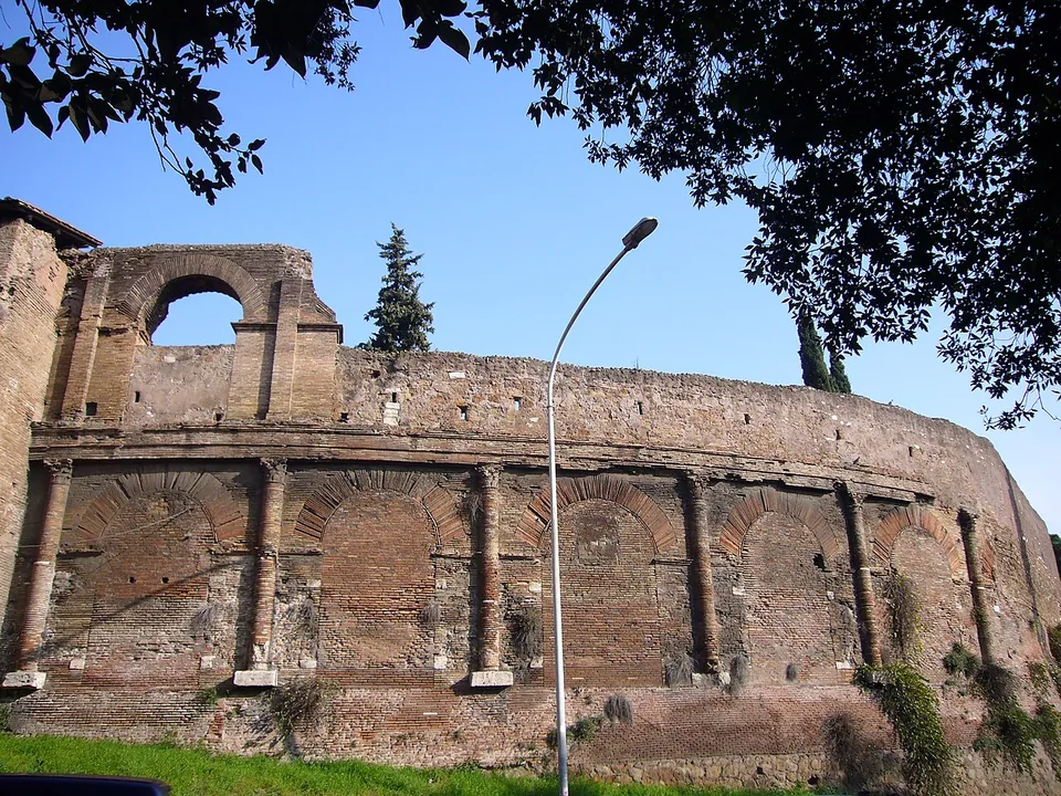 A long curved wall of red brick seen from the grass below, its lower storey a row of bricked-up arches between brick half-columns standing on stone bases, a moulded band above, and one open arch of an upper storey rising at the left end against the sky.