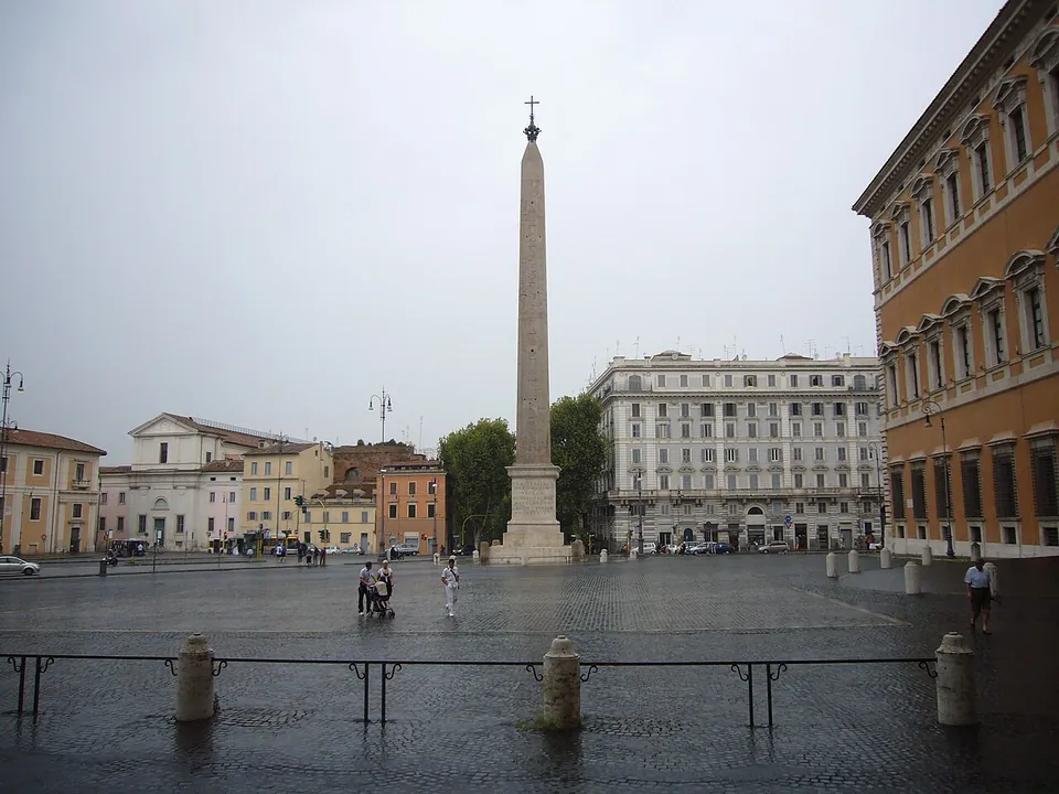 A wide cobbled square on an overcast day, a tall granite obelisk topped by a cross standing at its far side, with rows of apartment blocks behind and the orange flank of a palace at the right edge.
