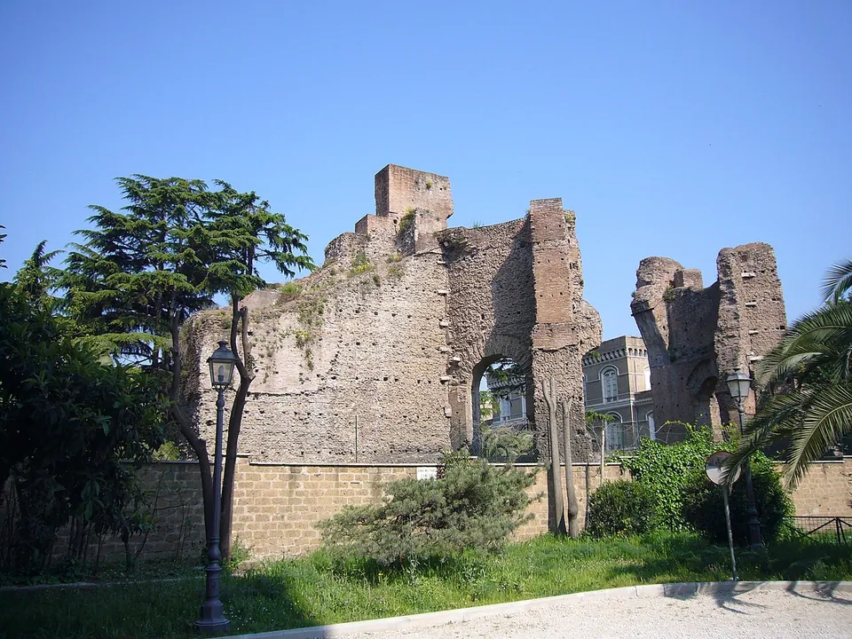 Tall ruined walls of brick and rubble with a large arched opening rising behind a modern boundary wall, with trees and a lamp post in front and a later building behind.
