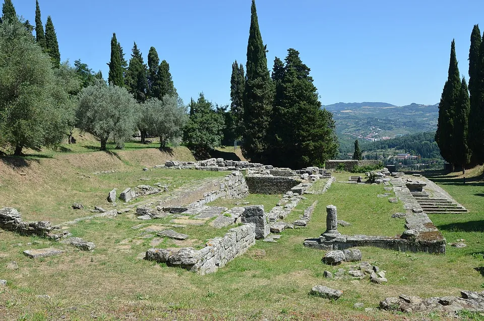 The low stone walls of a temple's podium and rooms laid out on a grassy terrace, a broad flight of steps on the right, cypresses and olive trees behind and wooded hills beyond.