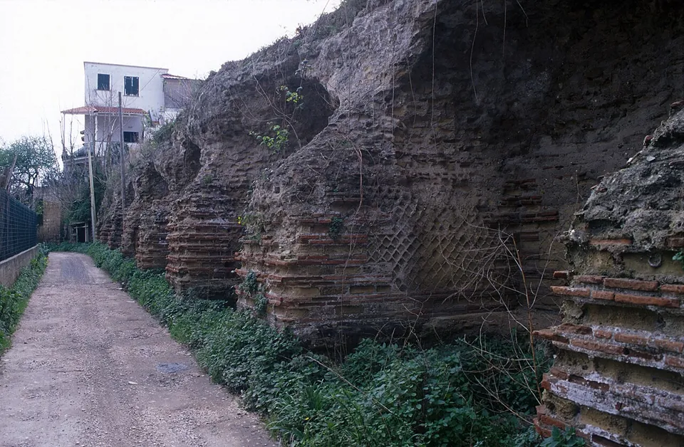 A lane running beside a high, overgrown wall of tufa and banded brick with diamond-pattern facing, set with buttresses, and a modern house at the far end.