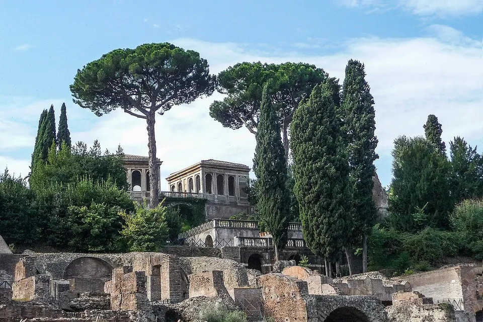 Umbrella pines and cypresses above a garden building with an open arcaded loggia on its upper floor, with low ruined brick walls in the foreground.