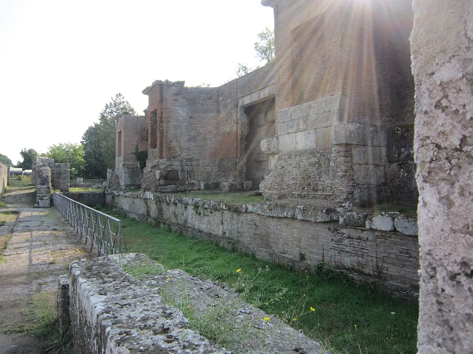 The ruined stage building of the theatre - tall walls of squared stone blocks and brick with a deep recess for a doorway - beside a paved path and a metal railing.