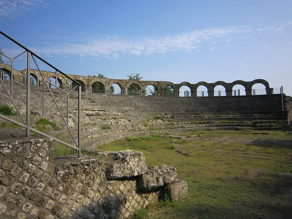 Rows of grey stone seating rising in a semicircle from a grassy floor, with a line of round arches on square piers running along the top and a metal railing in the foreground.