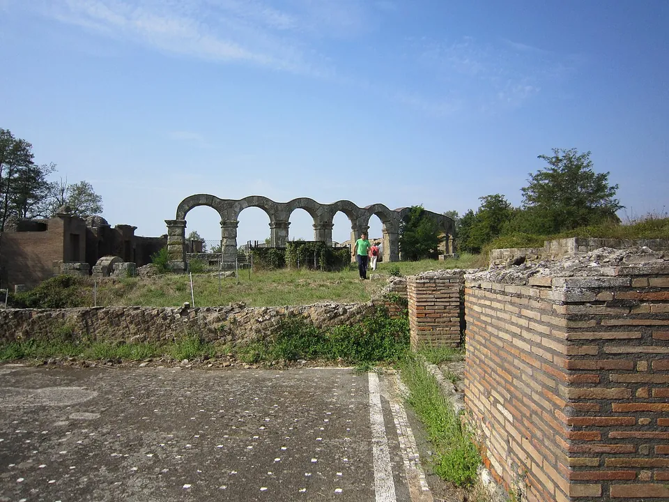 A dark mosaic floor dotted with small white squares, edged by a brick pier and low stone walls, with the theatre's arches and the stage building in the distance.