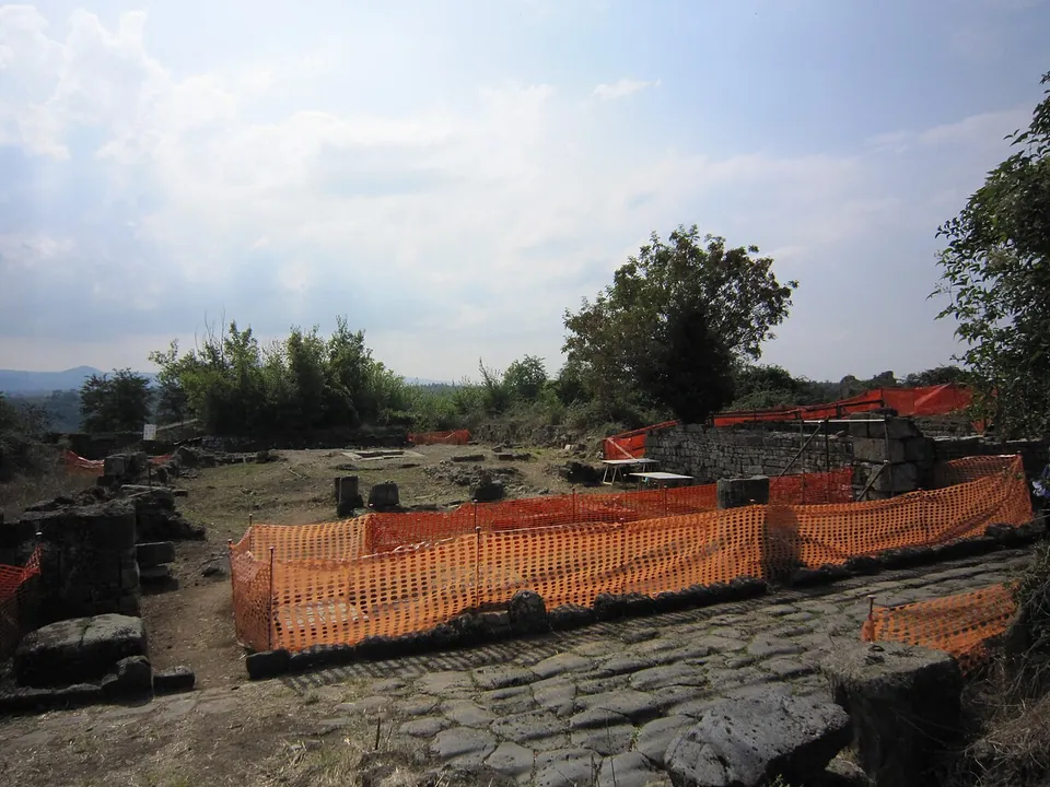 A street paved with large dark polygonal stones running past low excavated walls fenced off with orange mesh, with trees and distant hills beyond.