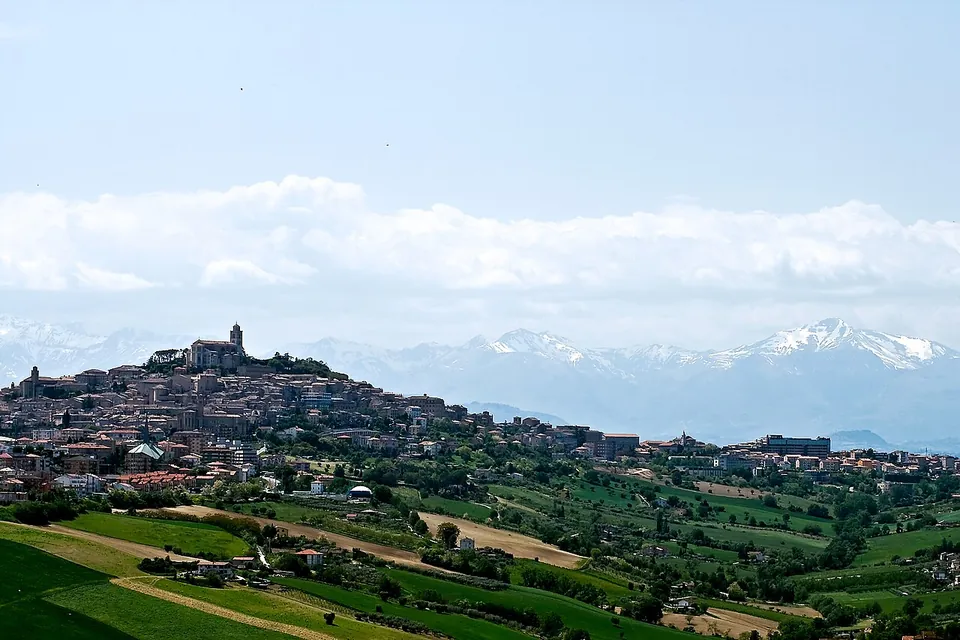 A hill town seen from across green farmland, its houses climbing to a summit crowned by a large building, with a range of snow-capped mountains on the horizon behind.