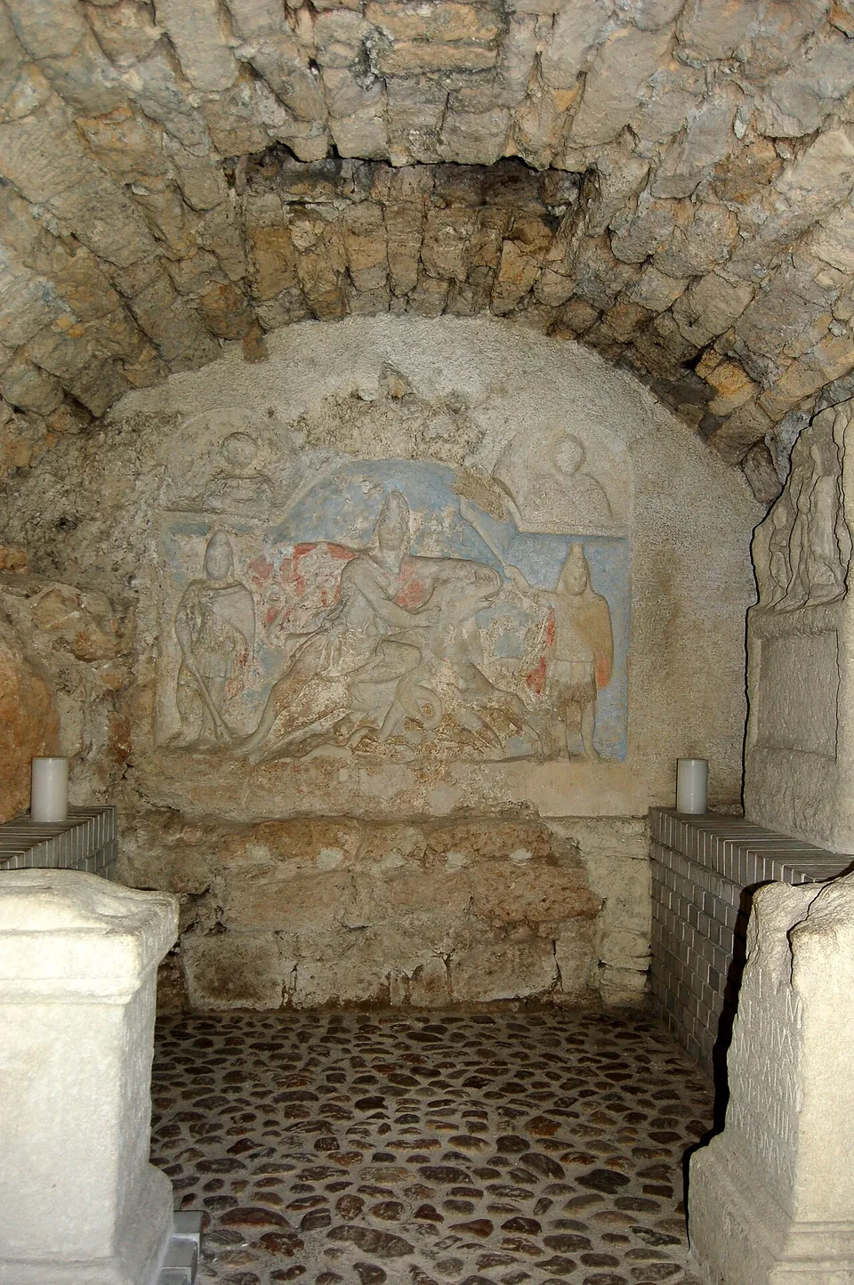 Inside a low vault of rough stone, a coloured relief on the end wall shows a figure in a cap pinning a bull with his knee, flanked by two standing torch-bearers and two busts above, with carved stone altars in the foreground on a cobbled floor.