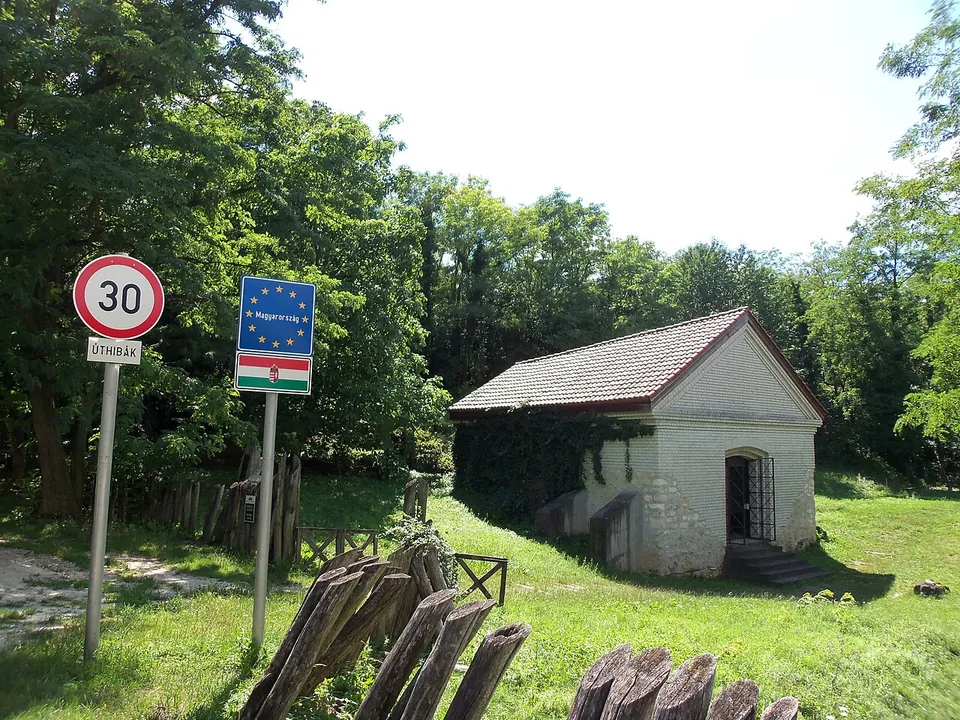 A small pale building with a tiled pitched roof and a barred door at the top of a few steps, among trees on a grassy slope, with a blue EU sign reading Magyarország and a 30 km/h speed-limit sign beside the road in front.