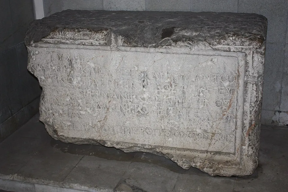 A large weathered block of white stone with a framed panel carved with several lines of Greek capital letters, set on a stone floor against a wall.