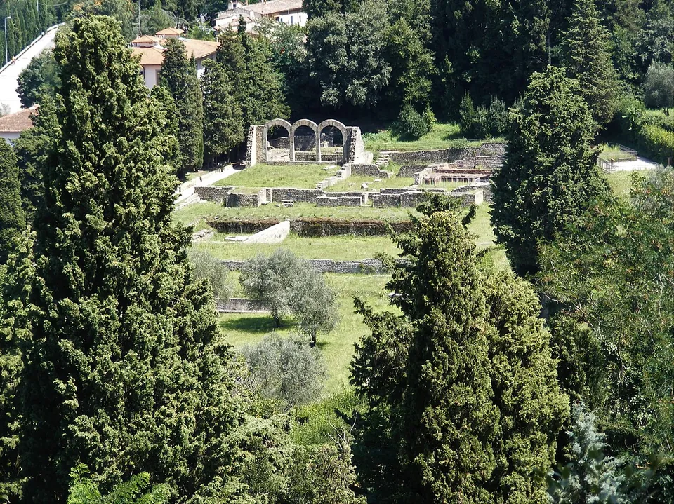 Looking down past tall cypresses onto a green hollow below the town, with low stone walls laid out on the lawns and, at the far side, three stone arches standing on their own.