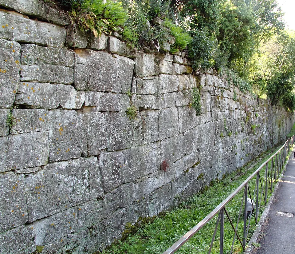 A tall wall of large, roughly squared grey sandstone blocks laid in courses, with plants growing along its top, beside a narrow footpath with an iron railing.