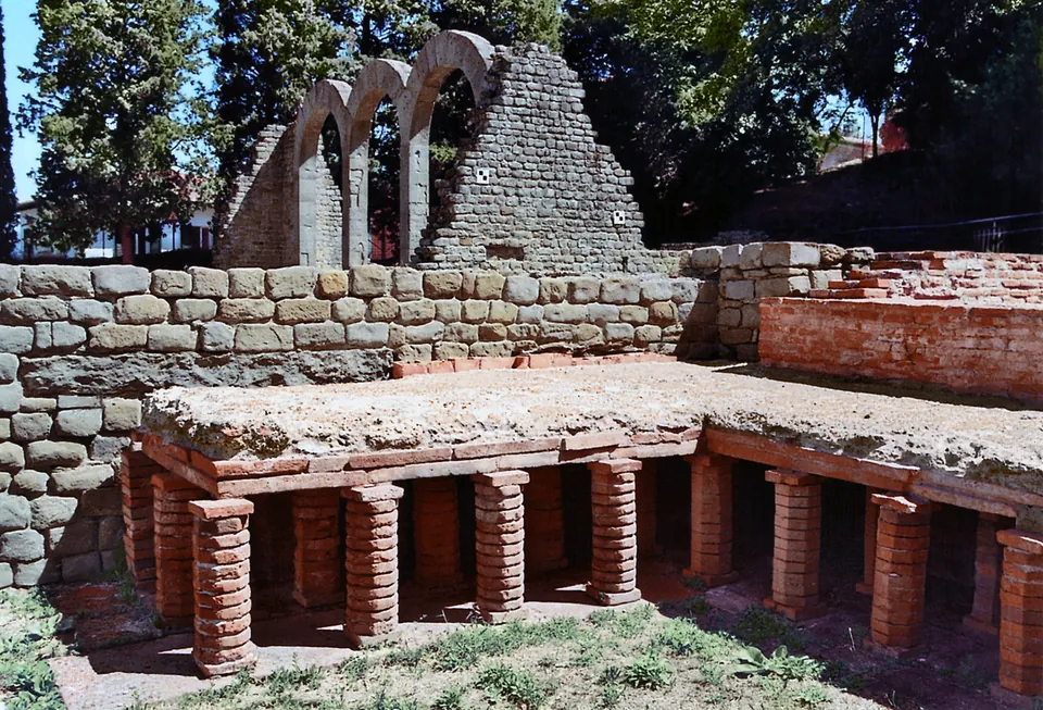Rows of short round brick pillars carrying a raised floor slab, set against a low wall of stone blocks, with the three stone arches rising behind.