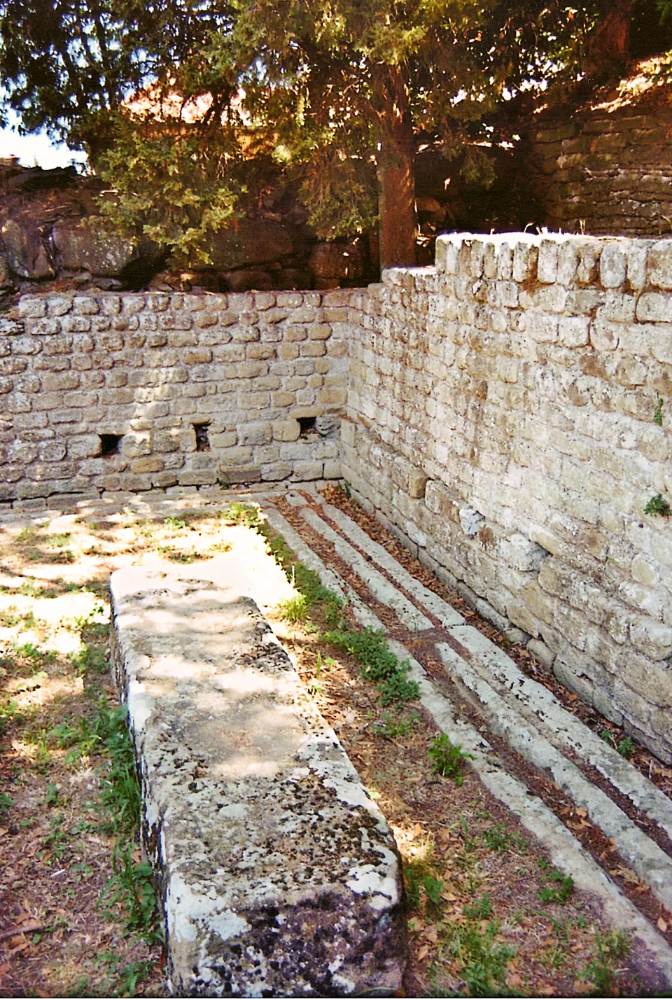 A small walled room of squared stone blocks, open to the sky, with a long stone block on the ground in front and worn stone edging along the foot of the right-hand wall.