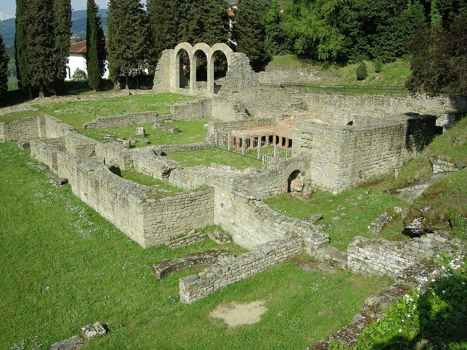 The baths seen from the slope above, a maze of low rectangular stone walls and grassy rooms, with a patch of red brick pillars in the middle and three stone arches standing at the far end among cypresses.