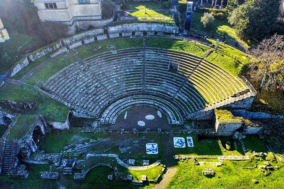 The semicircle of stone seating of the theatre seen from the air, its tiers divided by narrow stairways, with the orchestra and the low foundations of the stage building in front and lawns round about.