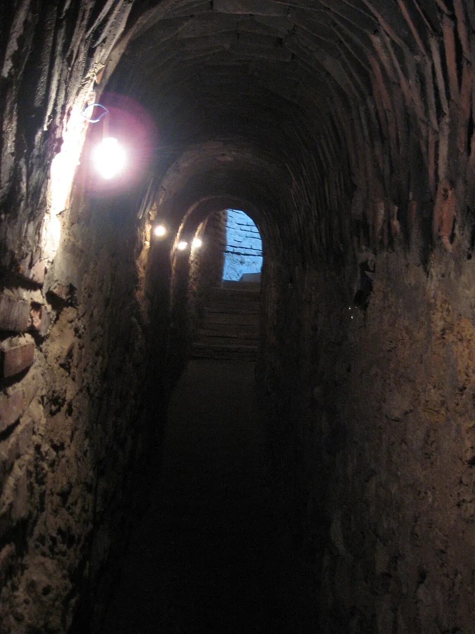 A narrow vaulted passage of old brick and stone, lit by lamps on the wall, running down steps towards daylight at the far end.
