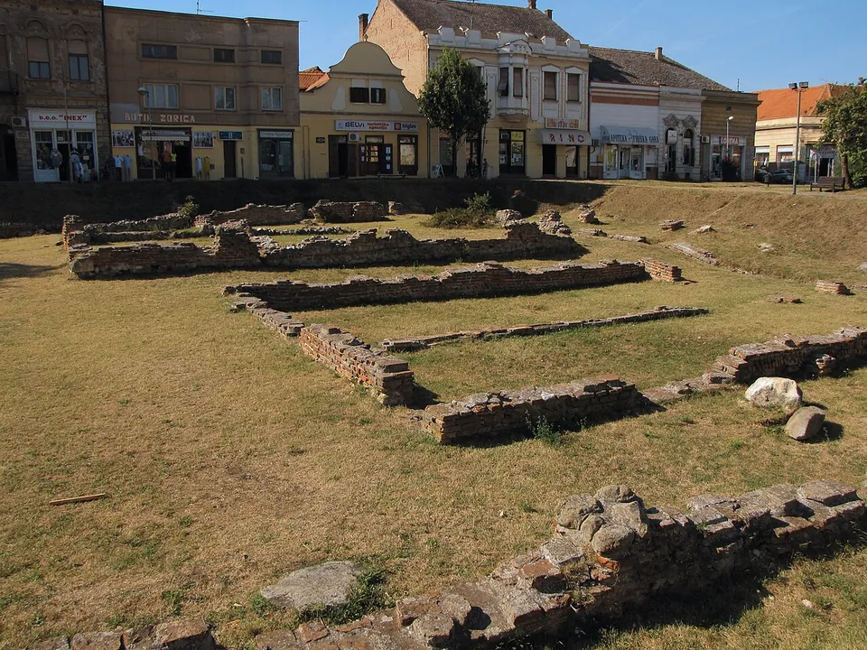 Low foundation walls of stone and brick mark out several rooms on a sunken lawn, with a grassy bank rising to a street of two-storey shopfronts behind.