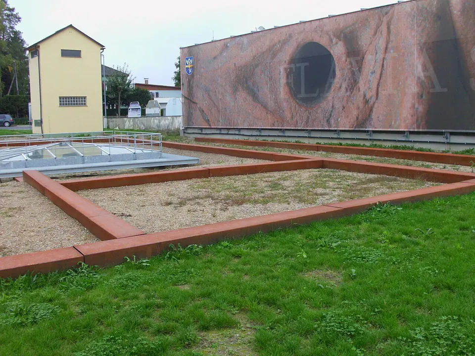 The outline of a building marked by rust-coloured metal edging on gravel in a grassy field, with a glass shelter and a large printed wall behind.