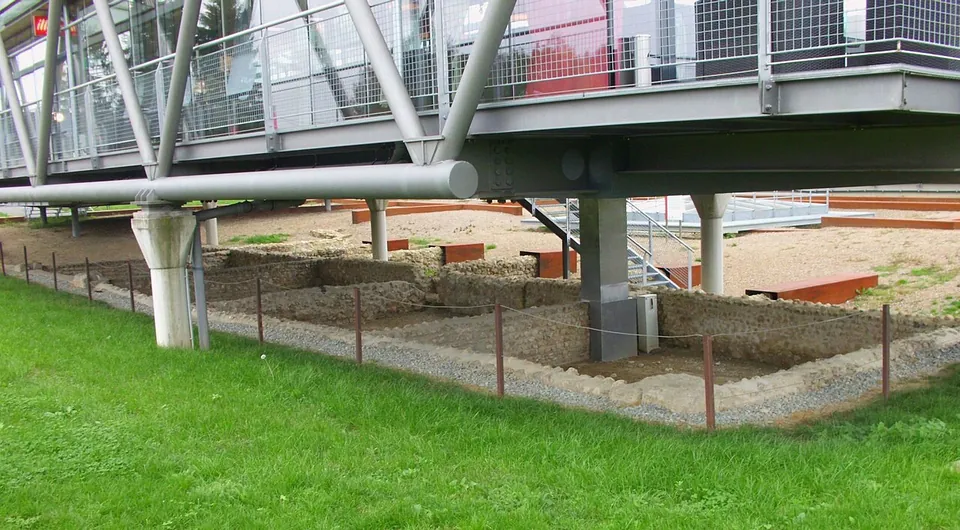 Low foundation walls inside a small fenced enclosure on a lawn, beneath the steel supports of a raised building.