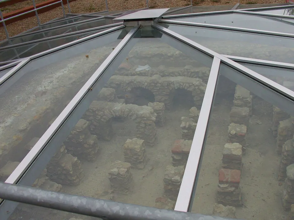 Looking down through a glass roof onto excavated walls of rubble stone and rows of small square brick pillars that once held up a heated floor.