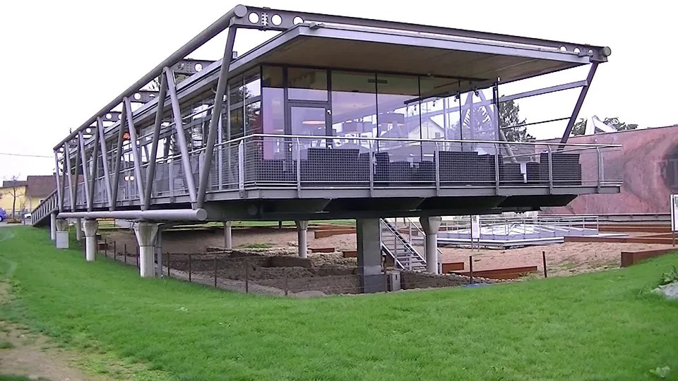 A modern glass-walled pavilion raised on steel stilts above a lawn, with fenced excavated ground beneath it.
