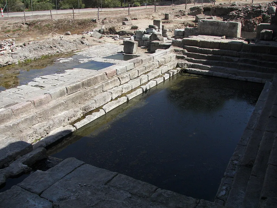 A rectangular pool lined with long stone blocks laid in steps, holding dark water, with ruined walls and loose blocks beyond it under a bright sky.