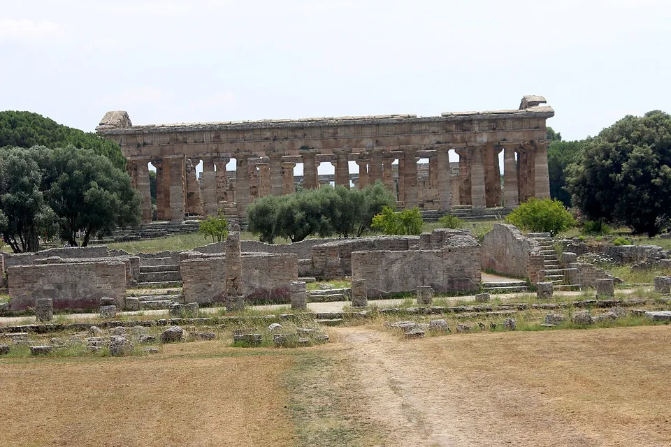 Low ruined walls of squared stone blocks and a few steps across a dry, grassy field, with a long Doric temple colonnade standing beyond them among trees.