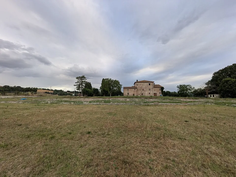 A wide flat field of dry grass with faint lines of low stonework at its far edge, a stone farmhouse and trees behind, under a cloudy sky.