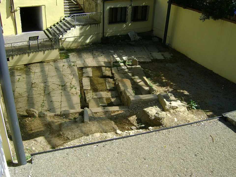 A sunken patch of large, uneven stone slabs and a few squared blocks, edged with gravel and closed in by the walls of modern buildings, with a flight of steps behind.