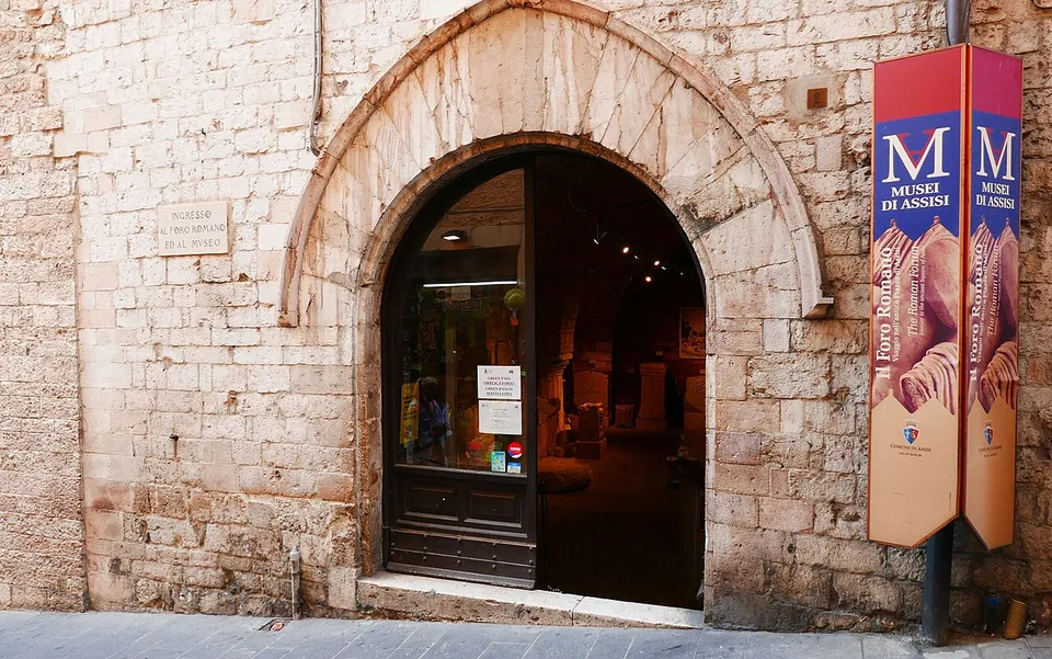A pointed stone archway in a wall of pale stone blocks, with a glazed door into a lit room, and a red banner beside it reading "Musei di Assisi" and "Il Foro Romano".