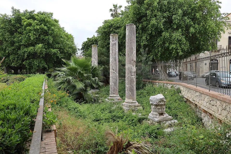 Three tall, slender marble column shafts standing among shrubs and palms in a sunken garden bed, with a carved stone block in front and a railing and parked cars along the street behind.