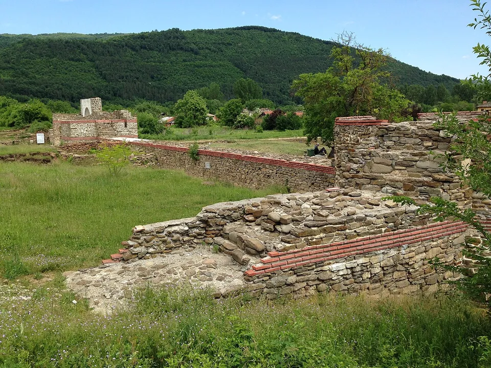 Restored walls of stone with brick courses crossing a grassy field, a small tower in the distance, and steep wooded mountains rising behind.