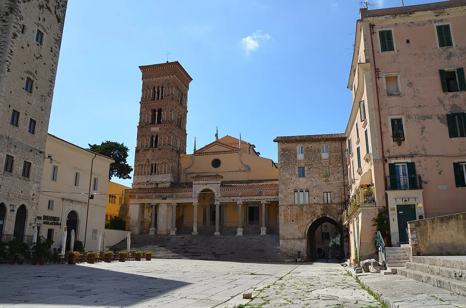A square paved with large worn stone slabs, closed by a church front with a columned porch at the top of a flight of steps and a tall brick bell tower, between old stone houses.